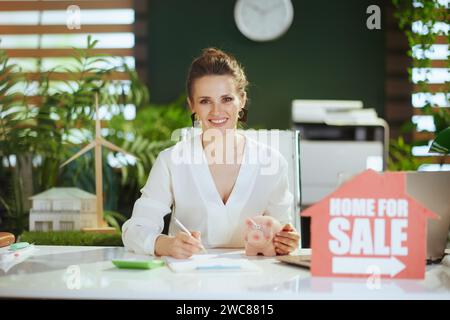 Umweltfreundliches Immobiliengeschäft. Lächelnde moderne 40 Jahre alte Geschäftsfrau in modernem grünen Büro in weißer Bluse mit Haus zum Verkauf Schild, Schweinebank, Calcu Stockfoto