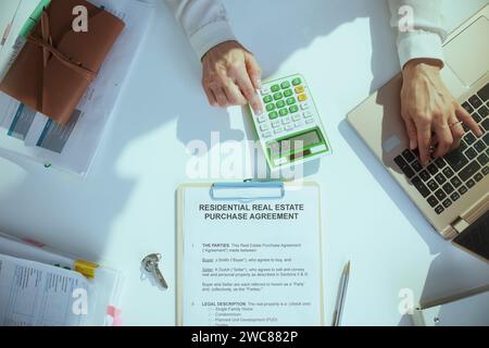 Es ist Zeit, weiterzumachen. Oberansicht einer Immobilienmaklerin im grünen Büro in weißer Bluse mit Rechner, Klemmbrett und Dokumentenarbeit am Laptop. Stockfoto