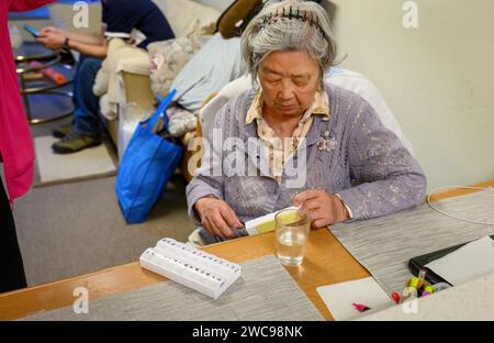Eine ältere Frau, die zu Hause ihre Medikamente in der Pillenbox überprüft. Familienmitglieder im Hintergrund. Kalifornien. Stockfoto