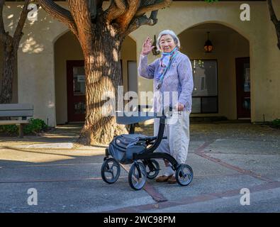 Eine ältere Frau mit einer Gesichtsmaske unter dem Kinn winkt zur Kamera und geht mit einem Mobilitätsläufer im Ruhestandort draußen. Stockfoto