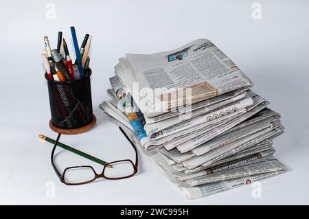 Stapel von Zeitungen, die gelesen werden müssen, um Informationen über Geschäfts- und Finanznachrichten zu erhalten, sowie Schreibwaren und Brillen auf dem Tisch Stockfoto