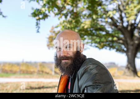 Ein Mann mit einem verdrehten langen Bart, der bei sonnigem Wetter lächelt. Er ist draußen, kahl und trägt eine Bomberjacke Stockfoto