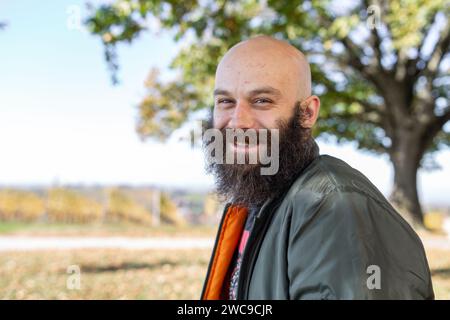 Ein Mann mit einem verdrehten langen Bart, der bei sonnigem Wetter lächelt. Er ist draußen, kahl und trägt eine Bomberjacke Stockfoto