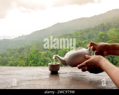 Person Hände halten keramische Teekanne gießen heißen Kräutertee in einer kleinen Tasse auf Holztisch auf natürlichem grünen Bergblick Landschaft Hintergrund mit Sonne Stockfoto