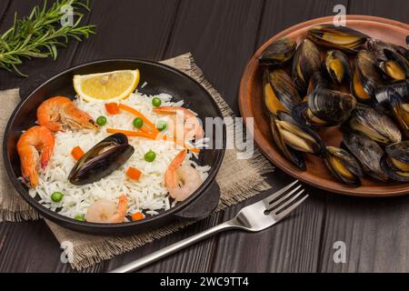 Garnelen und Muscheln mit Reis in der Pfanne. Muscheln auf dem Teller. Knoblauch, Zitrone und Rosmarin auf dem Tisch. Dunkler Holzhintergrund. Draufsicht Stockfoto