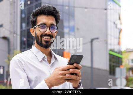 Lächelnder bärtiger Mann mit einer Brille, der in urbaner Umgebung ein Smartphone verwendet und Konnektivität, Freude und modernen Lebensstil vermittelt Stockfoto