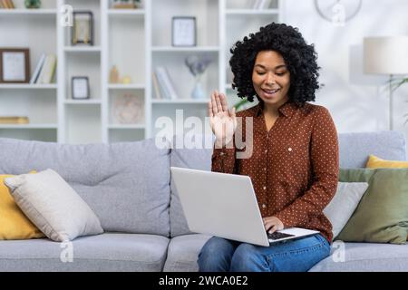 Lächelnde afroamerikanische junge Frau sitzt zu Hause auf dem Sofa, spricht per Laptop per Videoanruf, winkt und grüßt vor der Kamera. Stockfoto