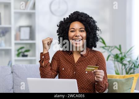 Porträt einer Afroamerikanerin, die einen Laptop hält, während sie zu Hause auf der Couch sitzt, eine Kreditkarte in der Hand hält und ihren Erfolg an der Kamera mit einer Siegesgeste genießt. Stockfoto