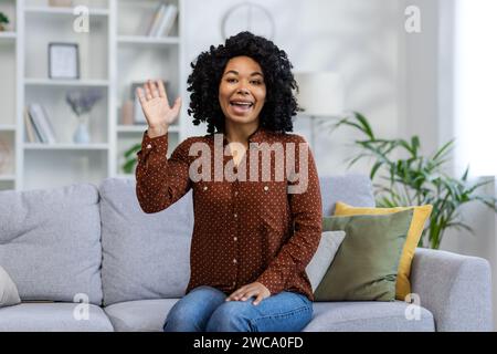 Porträt einer lächelnden jungen afroamerikanischen Frau, die zu Hause vor der Kamera auf der Couch sitzt, begrüßt und winkt. Stockfoto