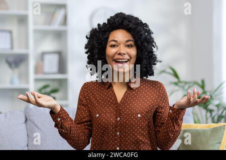Nahaufnahme eines Porträts einer jungen Afroamerikanerin, die zu Hause auf dem Sofa sitzt und die Kamera anlächelt, mit den Händen gestickt und in die Kamera blickt. Stockfoto