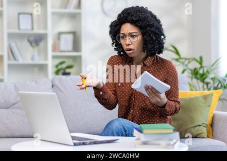 Besorgte junge afroamerikanische Studentin, die zu Hause durch Fernunterricht studiert, das Notebook in der Hand hält und frustriert über den Laptop-Bildschirm aussieht. Stockfoto