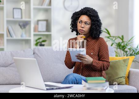 Nachdenkliche junge afroamerikanische Studentin, die zu Hause auf dem Sofa vor dem Laptop sitzt, ein Notebook mit Griff hält, eine Prüfung macht, Hausaufgaben macht. Stockfoto