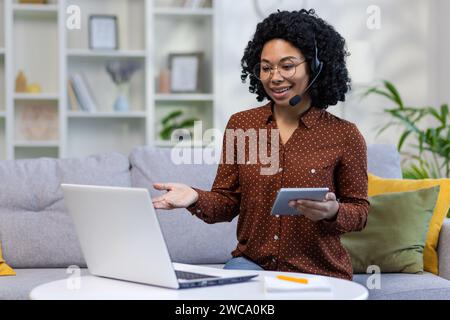 Eine junge afroamerikanische Lehrerin sitzt zu Hause in einem Headset vor einem Laptop, hält ein Tablet in der Hand und unterrichtet ferngesteuert. Stockfoto