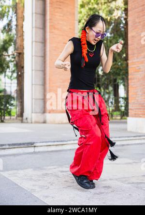 „Eine lebendige, dynamische Aufnahme, die eine junge Frau mit einem Flechtflecht einfängt, die auf der Straße tanzt, mit roten, sackigen Hosen und modischer lila Sonnenbrille.“ Stockfoto