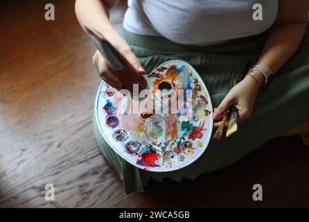 Hände einer Künstlerin, die schmutzige Palette mit verschiedenen Farben und Pinseln im Kunststudio hält Stockfoto