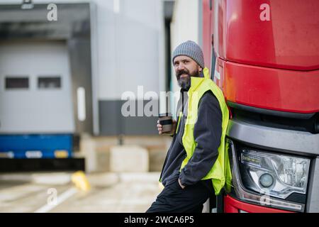 Lkw-Fahrer lehnt sich an den roten Lkw und trinkt Kaffee, wartet auf Lagerarbeiter. Stockfoto