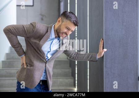 Erwachsener Mann in kluger Freizeitkleidung, der plötzliche Rückenschmerzen erlebt, vor Unwohlsein grimmt, sich drinnen an eine Wand lehnt. Stockfoto