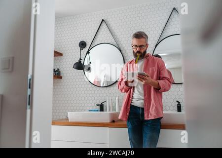 Ein Mann, der online einkaufen und sich im Badezimmer die Zähne putzt. Tablette halten, scrollen und kaufen, Zahnbürste im Mund. Stockfoto