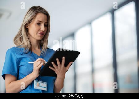 Porträt einer selbstbewussten Ärztin, die im Krankenhauskorridor steht. Schöne Krankenschwester mit blauen Schrubben und Klemmbrett in modernem Privat Stockfoto