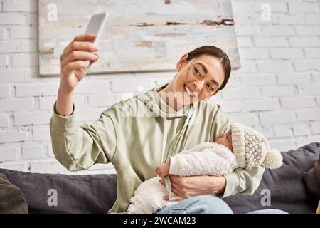 Fröhliche, schöne Mutter in lässiger Kleidung, die Selfie macht, während sie ihren neugeborenen Jungen hält Stockfoto