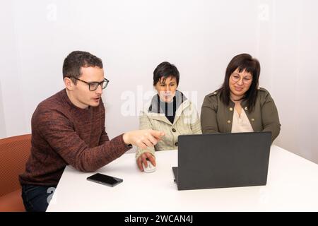 Drei Kollegen aus einem Büro, die sich in einem Besprechungsraum unterhalten. Sie sitzen an einem Tisch, auf dem sich ein Laptop und ein Smartphone befinden. Die Stockfoto