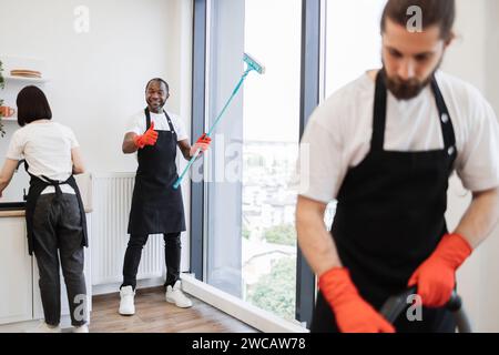 Konzentrieren Sie sich auf einen positiven afroamerikanischen Arbeiter, der Panoramafenster mit einem Wischtuch reinigt, in die Kamera schaut und den Daumen nach oben in einer modernen Wohnung in der Nähe von Multi zeigt Stockfoto