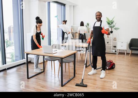 Afroamerikaner, der schwarze Schürze trägt und den Boden eines modernen Büros saugt. Stockfoto