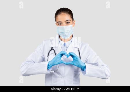 Mitfühlende europäische Ärztin in weißem Mantel und chirurgischer Maske, die mit ihren Händen eine Herzform macht Stockfoto