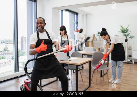 Glückliches multinationales Team von Leuten in schwarzen Schürzen, die modernes Büro reinigen. Stockfoto
