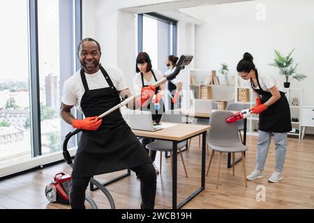 Glückliches multinationales Team von Leuten in schwarzen Schürzen, die modernes Büro reinigen. Stockfoto