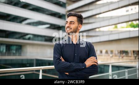 Ein lächelnder junger Geschäftsmann aus dem Nahen Osten steht im Freien selbstbewusst über seine Arme Stockfoto