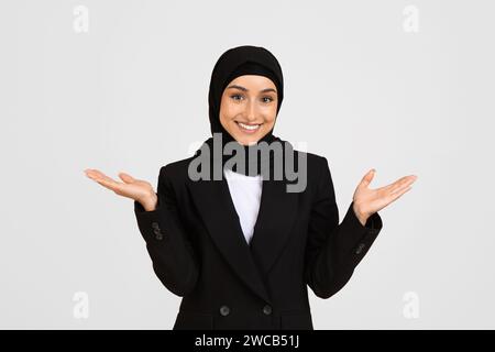 Glückliche junge muslimische Geschäftsfrau im Anzug und Hijab, die Hände mit offenen Händen hochhebt, sich freut und lächelt Stockfoto