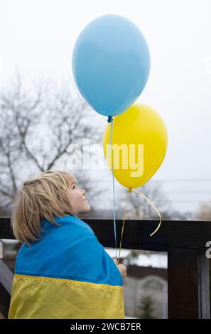 Junge in eine ukrainische Flagge gewickelt mit gelben und blauen Ballons. Glaube an den Sieg. Nostalgie für die Heimat Ukraine. Patriotisches Konzept. Stehen Sie bei der Ukraine Stockfoto