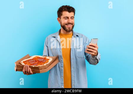 Mann mit Pizzakarton mit Smartphone, das über blauem Hintergrund steht Stockfoto