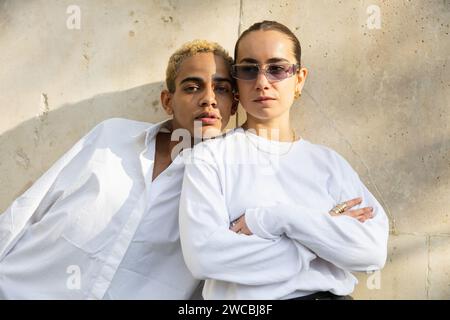 Eine junge Frau und ein Mann in knackiger weißer Kleidung teilen einen kurzen Moment und spiegeln ein Band der Freundschaft oder Verwandtschaft wider, das durch ein warmes, strukturiertes urbanes Wunderwerk hervorgehoben wird Stockfoto