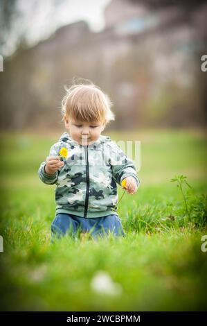 Glücklicher kleiner blonder Junge, der auf einem grünen Feld an einer schmackhaften Blume schnüffelt. Niedliches Kleinkind, das die Natur erkundet. Nahaufnahme Stockfoto