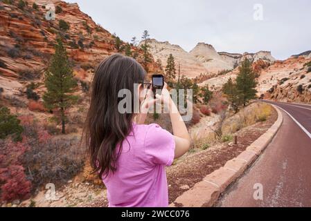 Ein Tourist macht ein Foto vom Straßenrand des Zion-Nationalparks. Es liegt in einem Tal, und viele der Büsche haben ihre Blätter verloren oder sind rot. Das g Stockfoto