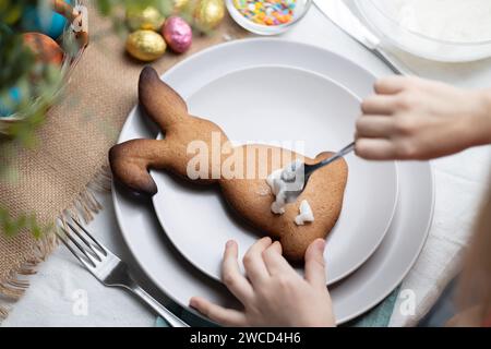 Nahaufnahme eines hasenförmigen Lebkuchenkekses. Kinderhände, die Kekse mit Glasur auf dem Teller dekorieren. Osterferien Stockfoto