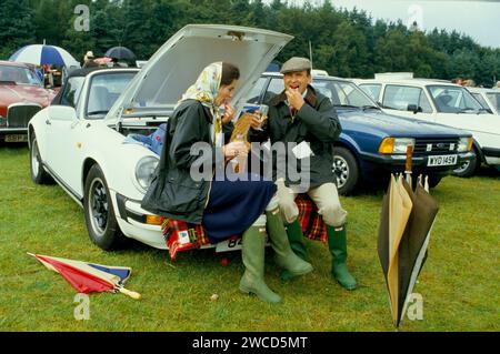 Das Paar Sloane Ranger genießt ein Picknick im Freien mit seinem Cabrio Porsche 911 SC Cabriolet. Sie ist auf einem Bucks Fizz, aber mit Henkell Trocken gemacht, einem deutschen Sekt, während er ihn ordentlich nimmt. Sie sind beide so gekleidet, wie es die Mode für den Anlass vorschreibt, in neuen, nicht matschigen grünen Hunter Wellington Boots und seinen und ihren blauen Burberry gewachsten Jacken, die damals absolut angesagt waren. Sie hat ein Hermes-Kopftuch angezogen, während er eine Tweed-Flatmütze trägt. Windsor, Berkshire, England. Juni 1985 1980. HOMER SYKES Stockfoto