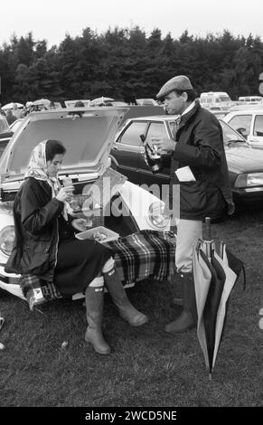 Polo Windsor Great Park, auf dem Gelände des Guards Polo Clubs genießt ein schickes Sloane Ranger-Paar ein Picknick im Freien von ihrem Cabriolet Porsche 911 SC. Sie ist auf einem Bucks Fizz, aber mit Henkell Trocken gemacht, einem deutschen Sekt, während er ihn ordentlich nimmt. Sie sind beide so gekleidet, wie es die Mode für den Anlass vorschreibt, in neuen, nicht matschigen grünen Hunter Wellington Boots und seinen und ihren blauen Burberry gewachsten Jacken, die damals absolut angesagt waren. Sie hat ein Hermes-Kopftuch angezogen, während er eine Tweed-Flatmütze trägt. Windsor, Berkshire, England. Juni 1985 1980. HOMER SYKES Stockfoto