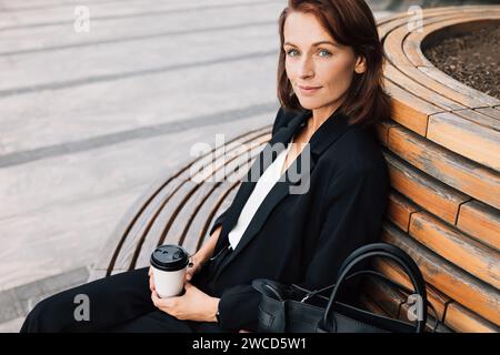 Lächelnde Geschäftsfrau mittleren Alters, die draußen auf einer Bank sitzt. Frau in Schwarz formell tragen entspannende Outdoor-Kleidung mit einer Tasse Kaffee. Stockfoto