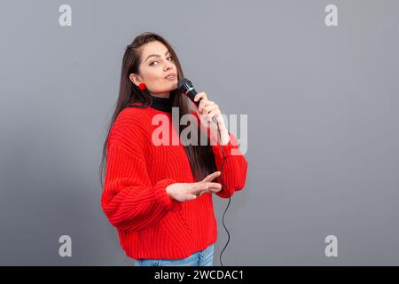 Junge, emotionale Brünette mit einem Mikrofon, das in einem roten Pullover gekleidet ist, singt Karaoke oder sagt eine Rede, isoliert auf Grau Stockfoto