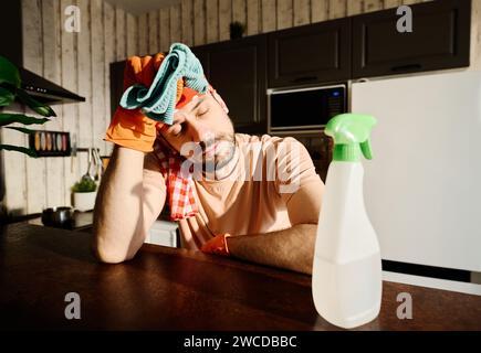 Erschöpfter Kerl in Gummihandschuhen und T-Shirt, der Hand mit blauem Staubtuch an Kopf hält, während er an der Küchenzeile mit Waschmittel steht Stockfoto