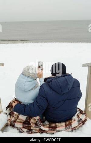 Glückliches älteres Paar, das im Winter auf der Treppe am Meer sitzt und ein Selfie macht. Vertikaler Rahmen. Stockfoto