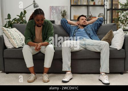 Junge verärgerte Afroamerikanerin, die neben ihrem gleichgültigen Ehemann auf der Couch sitzt und während des Streits beiseite schaut Stockfoto