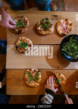Blick von oben nach unten auf die Hände, die mit Rucola garnieren und frisch gebackene kleine Pizzen auf einem Holztisch schneiden Stockfoto
