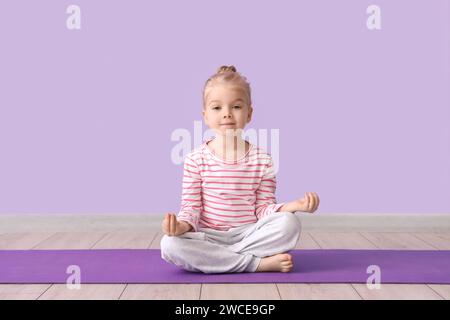 Niedliches kleines Mädchen, das in Lotusposition auf Yogamatte vor Fliederhintergrund sitzt Stockfoto