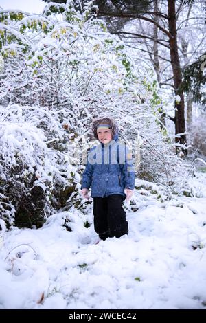 Kleines, schönes Mädchen in Winterkleidung, das allein in einem verschneiten Wald steht. Mädchen in Jacke mit Pelzhaube posiert im Winterwald. Sträucher A Stockfoto