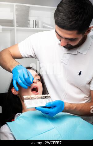 Eine Frau auf dem Zahnarztstuhl schaut auf seine Zähne und wählt die Farbe aus der Zahnpalette aus Stockfoto