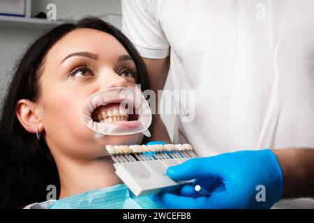 Eine Frau auf dem Zahnarztstuhl schaut auf seine Zähne und wählt die Farbe aus der Zahnpalette aus Stockfoto